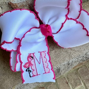 May include: White hair bow with pink scalloped edges and a pink flamingo embroidered with the letter "M" in the center.
