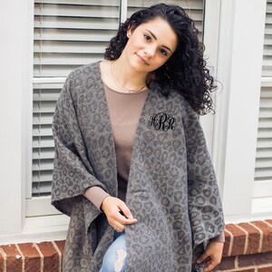 May include: A gray and brown leopard print poncho with fringe trim. The poncho has a monogrammed initial on the left side.