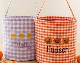 May include: Two fabric buckets with handles, one in lavender gingham with embroidered candy corn and the name Molly, the other in orange gingham with embroidered jack-o'-lanterns and the name Hudson, surrounded by small pumpkins and leaves.