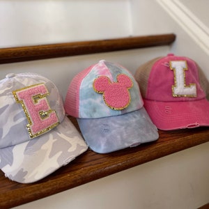 May include: Three pink and white baseball caps with different designs. The first cap has a camouflage pattern and a pink letter E with gold glitter. The second cap has a tie-dye pattern and a pink Mickey Mouse head with gold glitter. The third cap is solid pink with a white letter L with gold glitter.