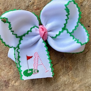 May include: White hair bow with green trim and a pink center. The bow has a white tag with green trim and an embroidered golf club and the letter "A".