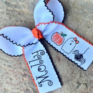 May include: White Halloween-themed hair bow with black scalloped edges and orange accents. The bow features embroidered images of an apple, ghost, and spider. The name "Molly" is embroidered on one of the ribbons.