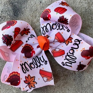 May include: A pink hair bow with a Thanksgiving theme. The bow is decorated with turkeys, pumpkins, pie slices, and leaves. The word "Molly" is written on the bow.