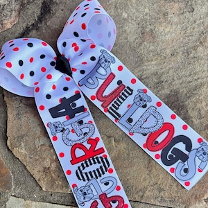 May include: A white hair bow with red and black polka dots. The bow features two streamers with the words "GEORGIA BULLDOGS" in black and red lettering, along with bulldog graphics. The bow is likely a fan accessory.