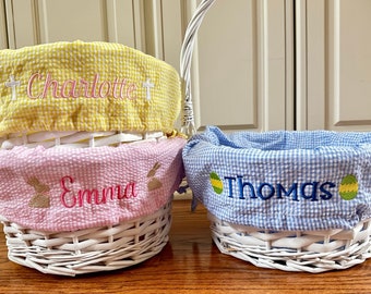 Może przedstawiać: Trzy białe wiklinowe koszyki wielkanocne z personalizowanymi podszewkami z tkaniny. Żółta podszewka ma wyszyte imię "Charlotte", różowa podszewka ma wyszyte imię "Emma", a niebieska podszewka ma wyszyte imię "Thomas".