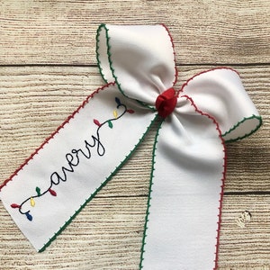 May include: White Christmas bow with green and red stitching around the edges. The bow has a string of colorful lights and the word "merry" embroidered on it.
