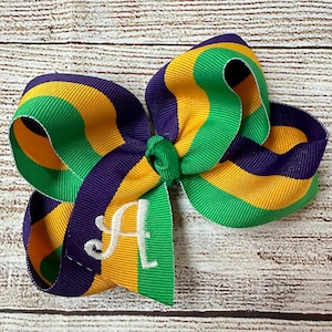 May include: A large, handmade hair bow in the colors of Mardi Gras: green, gold, and purple. The bow features a stitched white letter "A" on one of the tails. The bow is made of grosgrain ribbon.