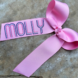 May include: A pink ribbon bow with the name "MOLLY" embroidered in colorful rainbow lettering.