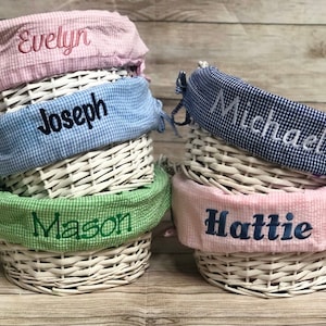 Pu&ograve; includere: Quattro cesti di vimini bianchi con fodere in tessuto. Le fodere sono rosa, blu, verde e vichy blu e bianco. Ogni fodera ha un nome ricamato: Evelyn, Joseph, Mason e Hattie.