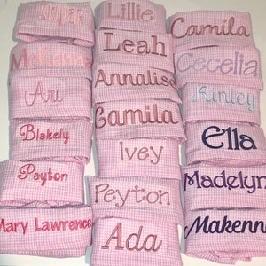 Pu&ograve; includere: Una pila di tessuto gingham rosa e bianco con vari nomi ricamati in diversi colori. I nomi includono Skylar, Lillie, Camila, McKenna, Leah, Cecelia, Ari, Annalisa, Finley, Camilla, Blakely, Ella, Ivey, Payton, Madelyn, Peyton, Mary Lawrence, Ada e Makenna.