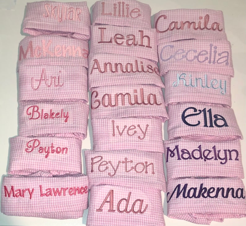 Pu&ograve; includere: Una pila di tessuto gingham rosa e bianco con vari nomi ricamati in diversi colori. I nomi includono Skylar, Lillie, Camila, McKenna, Leah, Cecelia, Ari, Annalisa, Finley, Camilla, Blakely, Ella, Ivey, Payton, Madelyn, Peyton, Mary Lawrence, Ada e Makenna.