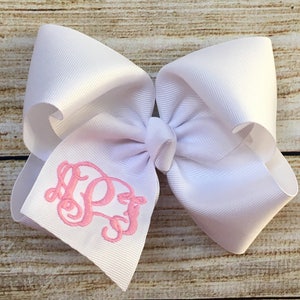 May include: A large, white hair bow with a pink monogram. The bow is made of a wide, ribbed fabric and features a three-letter monogram embroidered in a delicate pink thread. The bow is tied in the center.