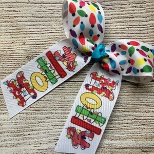 May include: A white hair bow with a red, green, yellow, and blue "HOLIDAY" design with colorful light bulbs. The bow is tied with a turquoise ribbon.
