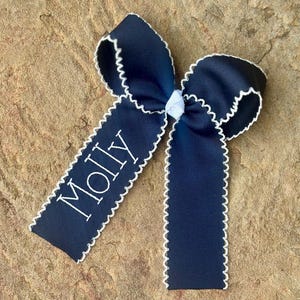 May include: Navy blue hair bow with white trim and the name "Molly" embroidered in white. The bow is made of a textured ribbon and has a classic bow shape. The bow is on a textured, tan surface.