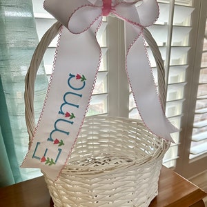 Puede incluir: Cesta de Pascua blanca tejida con un gran lazo de cinta blanca y una cinta personalizada. La cinta est&aacute; bordada con el nombre "Emma" en azul y presenta detalles florales. La cesta est&aacute; sobre una superficie de madera.