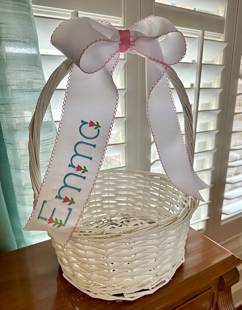 Puede incluir: Cesta de Pascua blanca tejida con un gran lazo de cinta blanca y una cinta personalizada. La cinta est&aacute; bordada con el nombre "Emma" en azul y presenta detalles florales. La cesta est&aacute; sobre una superficie de madera.