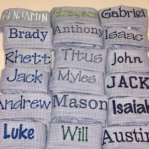 Pu&ograve; includere: Una pila di quadrati di tessuto gingham blu e bianco con vari nomi ricamati in filo blu. I nomi includono Benjamin, Grayson, Gabriel, Brady, Anthony, Isaac, Rhett, Titus, John, Jack, Myles, JACK, Andrew, Mason, Isaiah, Luke, Will e Austin.