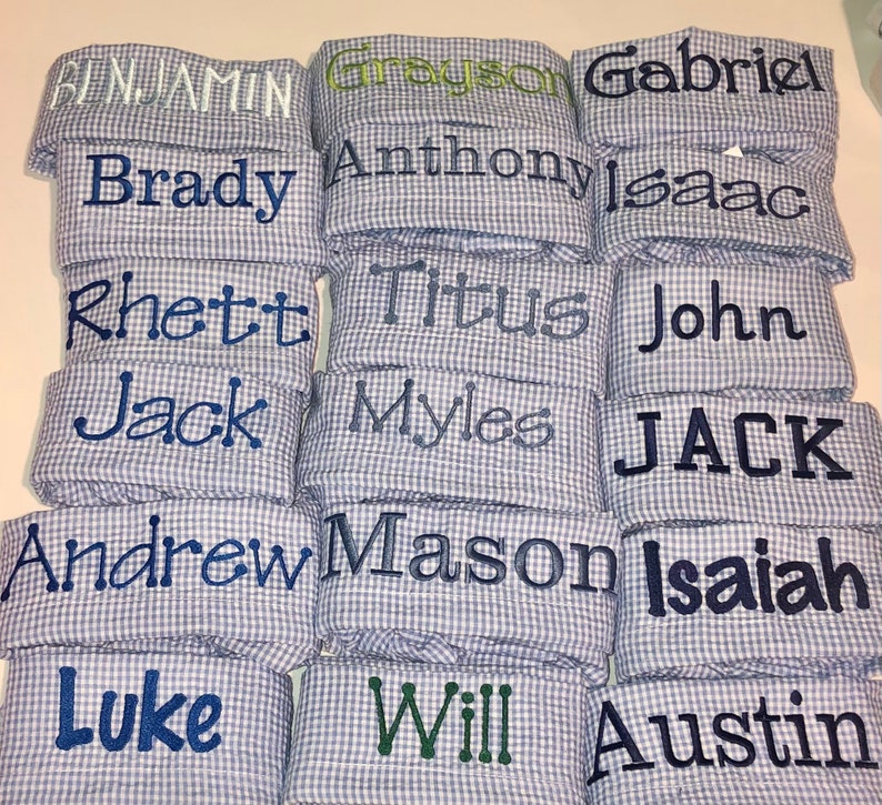 Pu&ograve; includere: Una pila di quadrati di tessuto gingham blu e bianco con vari nomi ricamati in filo blu. I nomi includono Benjamin, Grayson, Gabriel, Brady, Anthony, Isaac, Rhett, Titus, John, Jack, Myles, JACK, Andrew, Mason, Isaiah, Luke, Will e Austin.