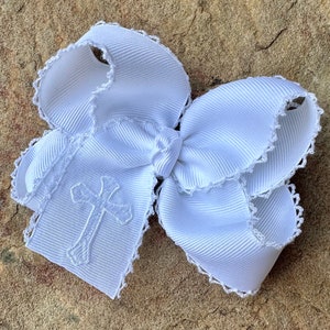 May include: White satin hair bow with a white embroidered cross design. The bow has a scalloped edge.