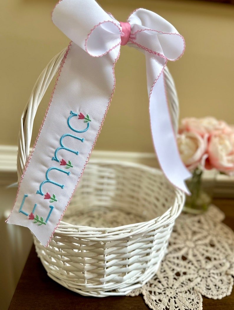 Puede incluir: Cesta blanca tejida con un gran lazo de cinta blanca y una cinta personalizada. La cinta est&aacute; bordada con el nombre "Emma" en letras azul claro con detalles florales rosas. Ideal para Pascua o para regalar.