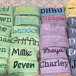 Pu&ograve; includere: Una pila di quadrati di tessuto gingham verde, blu, viola e giallo con nomi ricamati su ogni quadrato. I nomi includono David, Shawn, Jace, Thomas, Kyson, Chandler, Perrin, Reegan, Landon, Griffin, Millie, Deven, DHAU, Maya, Freya, Charley, Hannah, Nico, Ryan, Dhilhan, Emmett e Jovan.