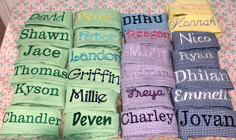 Pu&ograve; includere: Una pila di quadrati di tessuto gingham verde, blu, viola e giallo con nomi ricamati su ogni quadrato. I nomi includono David, Shawn, Jace, Thomas, Kyson, Chandler, Perrin, Reegan, Landon, Griffin, Millie, Deven, DHAU, Maya, Freya, Charley, Hannah, Nico, Ryan, Dhilhan, Emmett e Jovan.