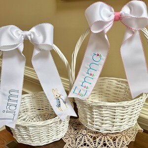 Puede incluir: Dos cestas de Pascua blancas de mimbre con grandes lazos de sat&eacute;n blanco y cintas personalizadas. Una cinta dice "Liam" en azul marino, la otra "Emma" en azul claro con detalles florales. Una cinta tiene un bordado de Peter Rabbit.