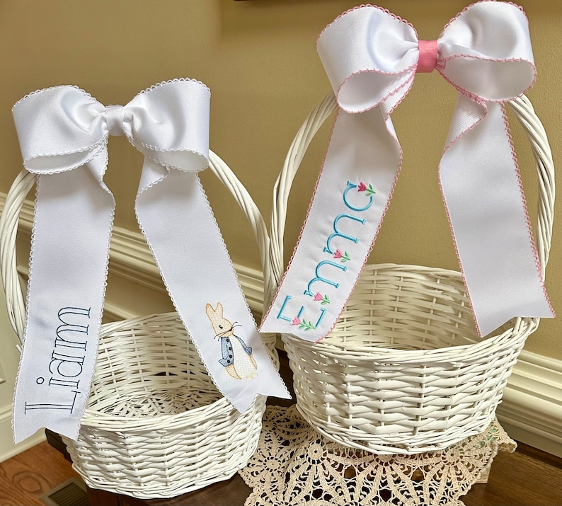 Puede incluir: Dos cestas de Pascua blancas de mimbre con grandes lazos de sat&eacute;n blanco y cintas personalizadas. Una cinta dice "Liam" en azul marino, la otra "Emma" en azul claro con detalles florales. Una cinta tiene un bordado de Peter Rabbit.