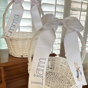 Puede incluir: Dos cestas de Pascua de mimbre blanco con lazos de cinta blancos. Una cinta dice "Emma" con detalles florales, la otra "Liam" con un bordado de Peter Rabbit. Las cintas tienen un borde festoneado.