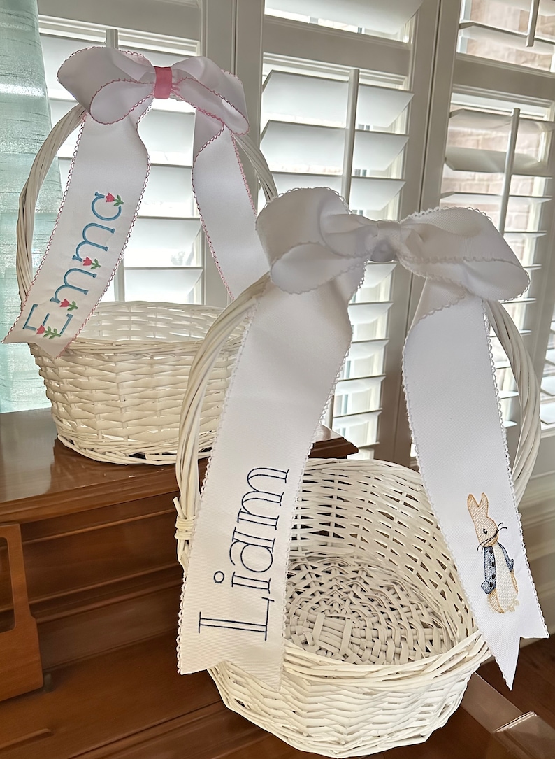 Puede incluir: Dos cestas de Pascua de mimbre blanco con lazos de cinta blancos. Una cinta dice "Emma" con detalles florales, la otra "Liam" con un bordado de Peter Rabbit. Las cintas tienen un borde festoneado.