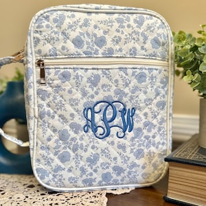 May include: A quilted, square-shaped bag with a floral pattern in light blue and white. The bag features a zipper closure, a handle, and a blue embroidered monogram. The bag is approximately 9 inches tall and 8 inches wide.