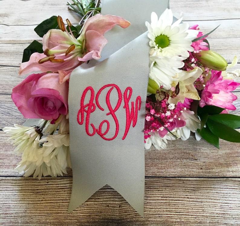 May include: A gray satin ribbon with a pink monogram "E.S.W." embroidered on it. The ribbon is tied around a bouquet of pink, white, and green flowers.