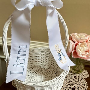 Puede incluir: Cesta de Pascua de mimbre blanco con un lazo de cinta blanca. La cinta tiene bordado el nombre "Liam" en azul marino y un dise&ntilde;o de Peter Rabbit. La cesta est&aacute; sobre una mesa con un tapete de encaje y un jarr&oacute;n con flores rosas.