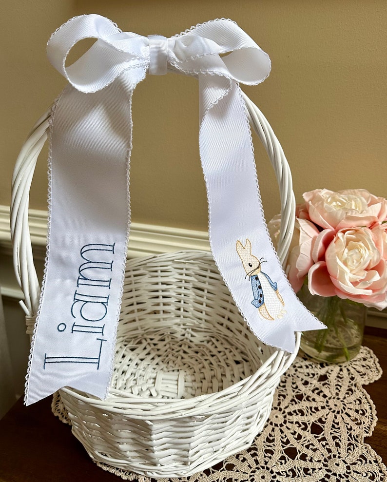 Puede incluir: Cesta de Pascua de mimbre blanco con un lazo de cinta blanca. La cinta tiene bordado el nombre "Liam" en azul marino y un dise&ntilde;o de Peter Rabbit. La cesta est&aacute; sobre una mesa con un tapete de encaje y un jarr&oacute;n con flores rosas.