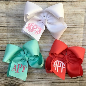 May include: Three large hair bows with different colored fabric and personalized monograms. The bows are white, teal, and coral.