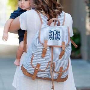 May include: A blue and white striped canvas backpack with brown leather accents and a large monogrammed initial "JRB" on the front flap.
