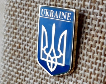 May include: A blue and gold enamel pin with the Ukrainian coat of arms, a trident, and the word "UKRAINE" above it.