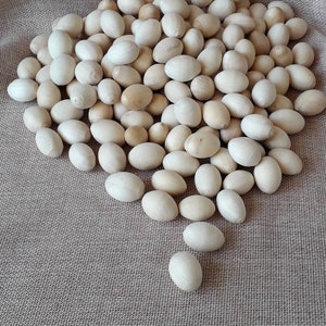 Op de afbeelding: Een grote stapel gladde, houten eieren in een lichtbeige kleur. De eieren zijn willekeurig gerangschikt op een jute oppervlak.