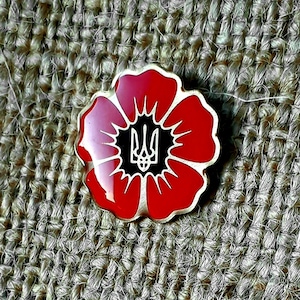 Może przedstawiać: Szpilka w kształcie czerwonego maku z złotym ukraińskim trójzębem w środku. Szpilka znajduje się na brązowym tle z płótna.