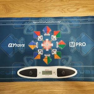 May include: A blue and white  'CUBE'  mat with a colorful design in the center. The mat has the words 'QYTOYS' and 'M PRO' on either side. A white digital scale with a black display reads '0.000'.