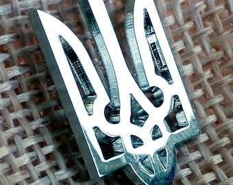 May include: A silver-colored metal trident, a symbol of Ukraine, is displayed on a woven fabric background.