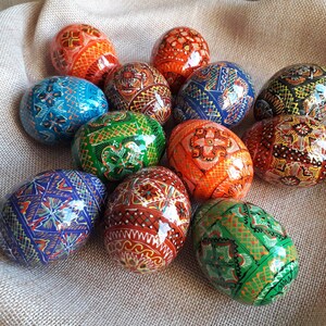 May include: A collection of twelve intricately painted Easter eggs, each featuring a unique geometric design. The eggs are displayed on a burlap background and showcase a variety of colors, including blue, green, orange, red, and yellow.