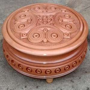 May include: A round wooden box with intricate carvings on the lid. The box is brown and has a glossy finish. It sits on three small feet.