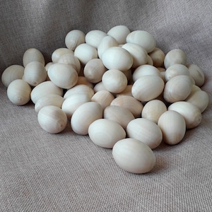 Op de afbeelding: Een stapel houten eieren in verschillende maten en vormen, allemaal geverfd in een natuurlijke houtkleur. De eieren zijn gerangschikt op een textuurrijke stoffen achtergrond.