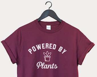 Puede incluir: Camiseta color burdeos con texto blanco que dice "Powered by Plants" y un pequeño icono de planta.
