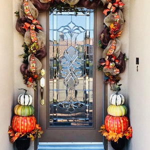 May include: A fall-themed door wreath with brown, orange, and green mesh, ribbons, and pumpkins. Two stacked pumpkins in a black planter are on either side of the door. The doormat says "Welcome".