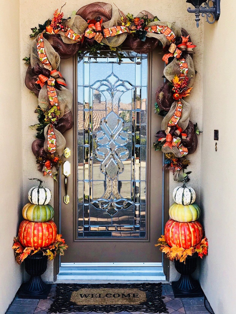 May include: A fall-themed door wreath with brown, orange, and green mesh, ribbons, and pumpkins. Two stacked pumpkins in a black planter are on either side of the door. The doormat says "Welcome".
