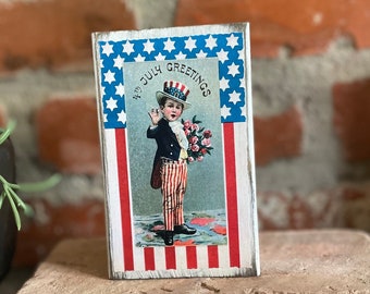 May include: A wooden sign with a red, white, and blue American flag design. The sign features a vintage image of a young boy dressed in a red, white, and blue outfit, holding a bouquet of flowers. The sign reads "4th of July Greetings".