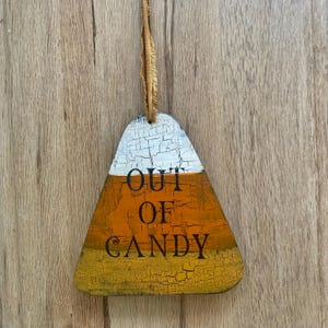 May include: Wooden candy corn-shaped decoration with the text "OUT OF CANDY". The top is white, the middle is orange, and the bottom is yellow. A brown string is attached for hanging.