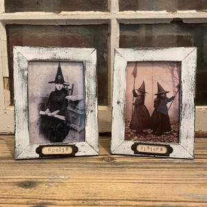 May include: Two white picture frames with black accents, each featuring a vintage image of a witch. The frame on the left has the text "Spells" on a small tag below the image. The frame on the right has the text "Sisters" on a small tag below the image.